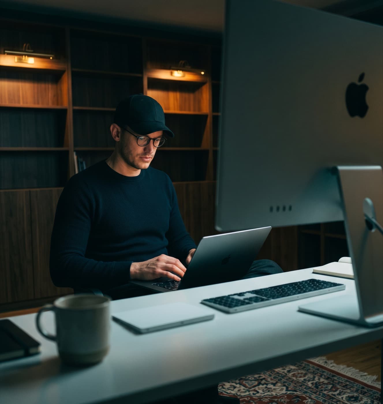 Mann mit schwarzer Kappe und Brille, der an einem Laptop arbeitet, in einem dunklen Raum mit Holzregalen.